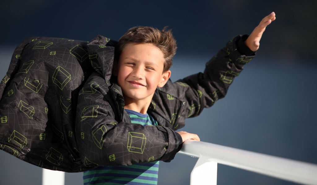 Веселый мальчик в куртке стоит на ветру в лучах солнца