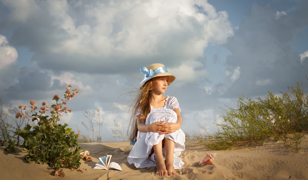 Маленькая девочка в белом платье сидит на песке 