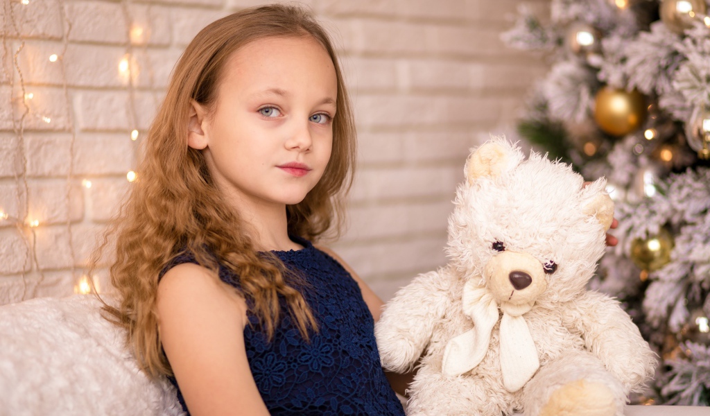 Маленькая девочка с мишкой сидит у наряженной ели 