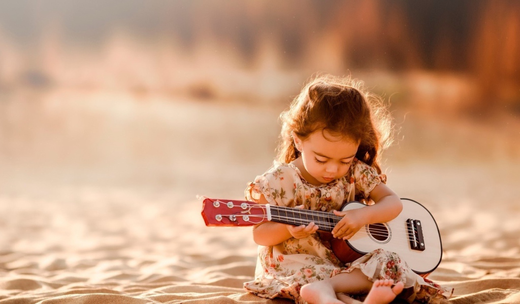 Маленькая девочка с гитарой сидит на песке 