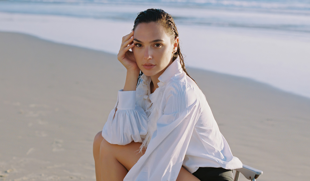 Актриса Галь Гадот в белой рубашке сидит на песке