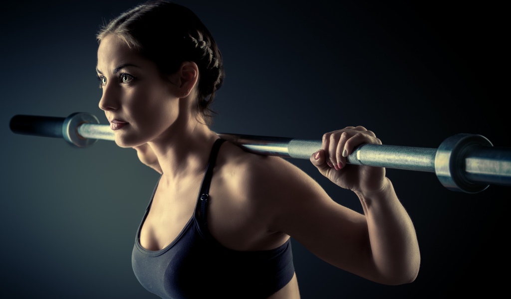 The sports girl is engaged with a barbell in the gym on a gray background