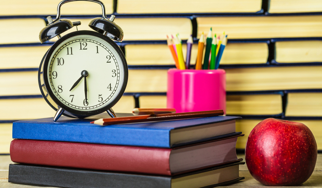 Будильник, книги, карандаши и яблоко на столе у стены 