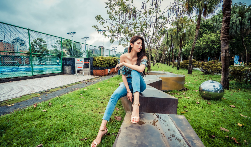 Девушка азиатка в джинсах сидит в парке 