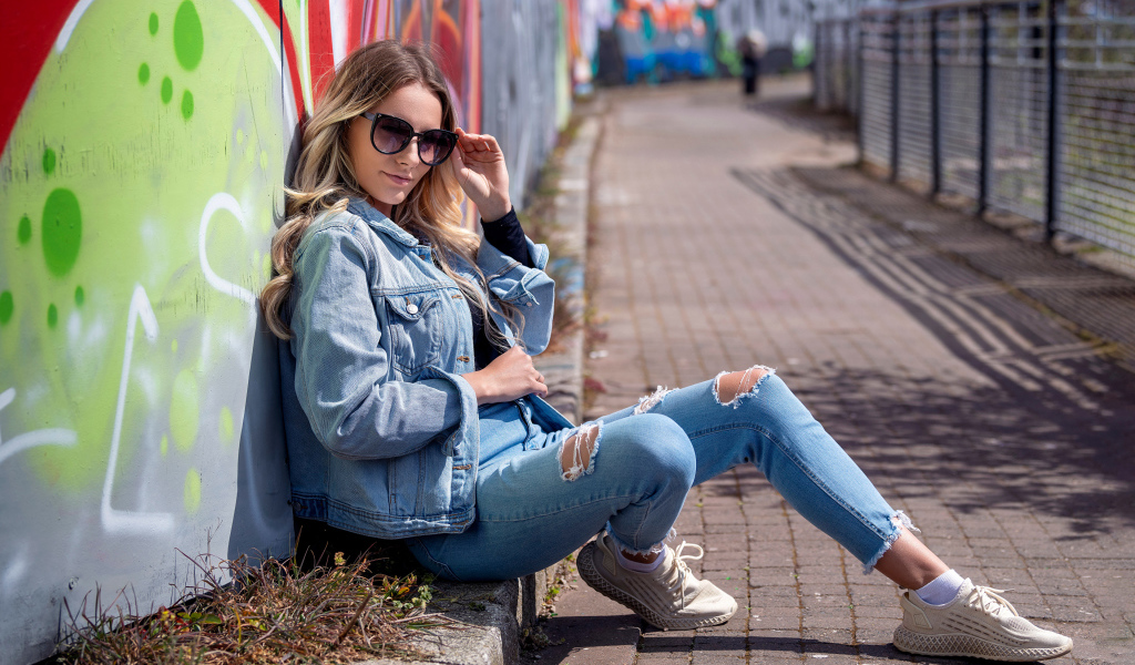 Красивая девушка в джинсовом костюме сидит у стены 