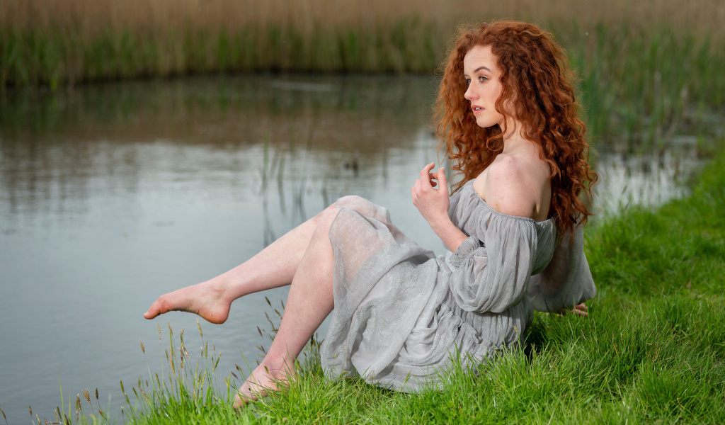 Красивая девушка с рыжими волосами сидит на берегу пруда 