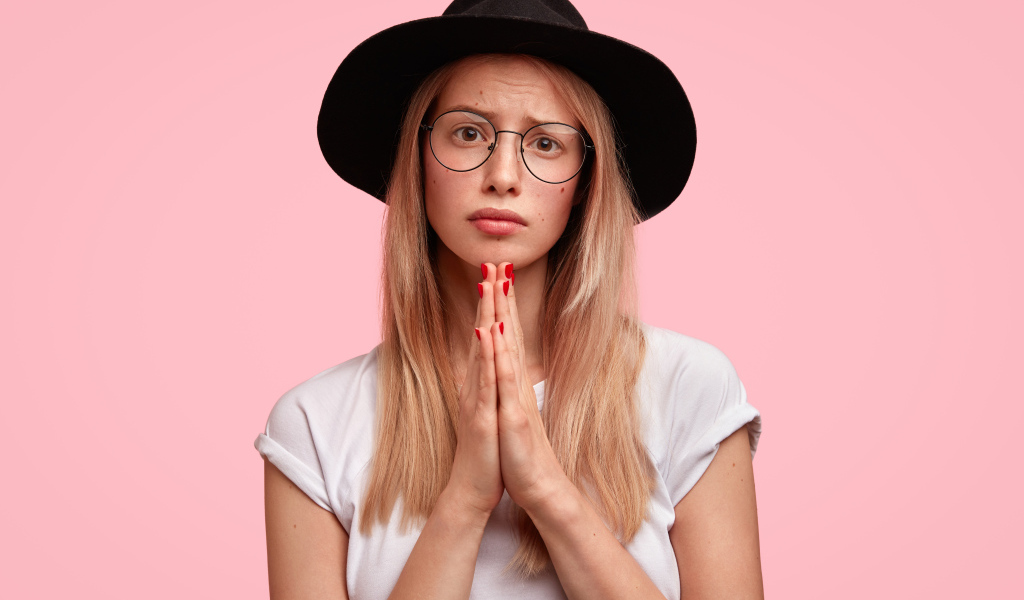 Девушка в черной шляпе на розовом фоне просит прощения