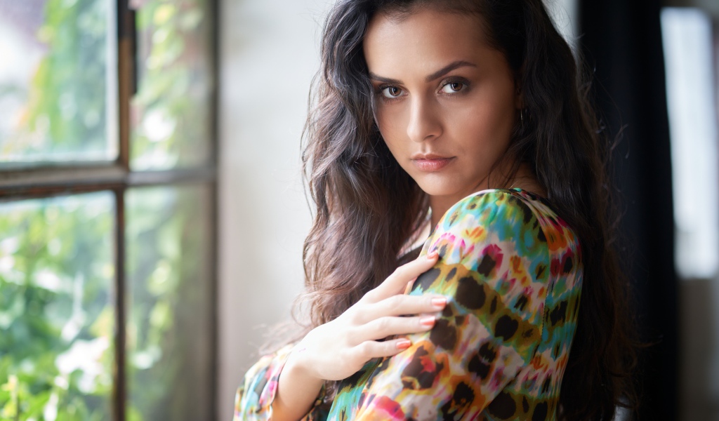 Длинноволосая кареглазая девушка стоит у окна