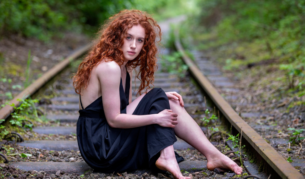 Рыжеволосая девушка в черном платье сидит на путях