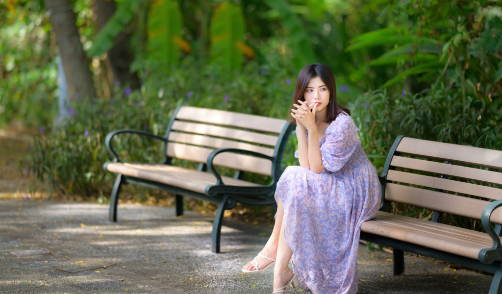 Грустная девушка азиатка в платье на лавке в парке 