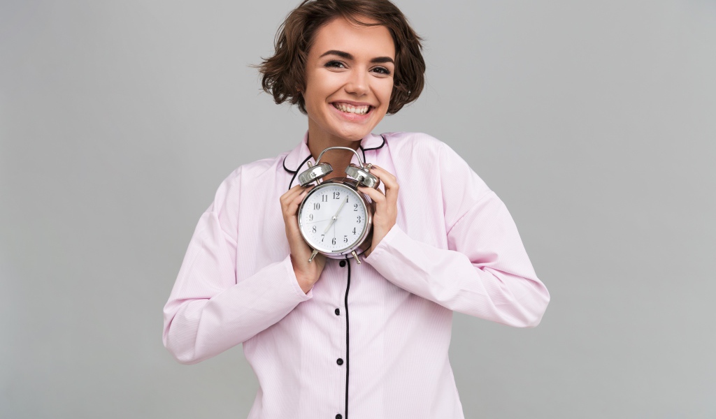 Улыбающаяся девушка в розовой рубашке с будильником в руках 