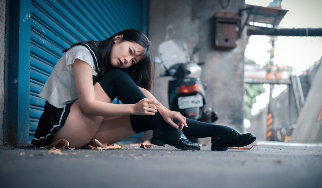 Молодая азиатка в черных чулках сидит у стены 