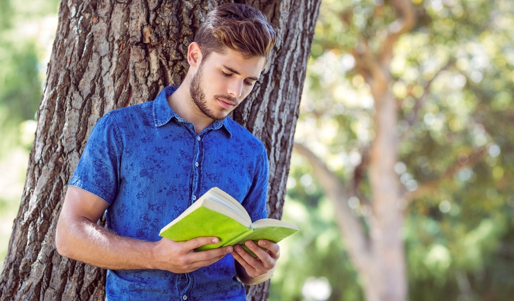 Красивый молодой мужчина стоит с книгой под деревом 