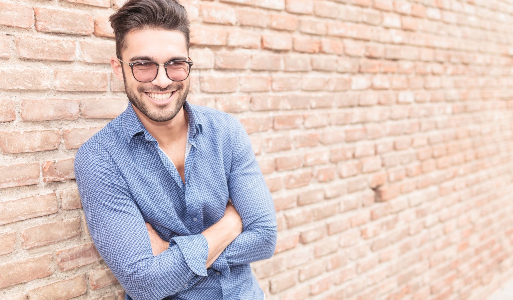 Улыбающийся мужчина в очках стоит у стены 