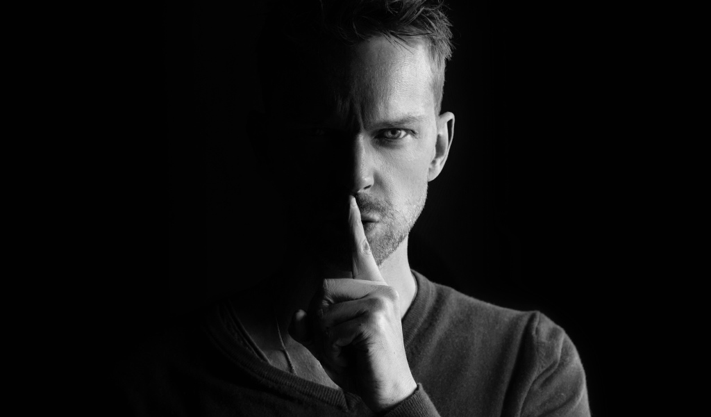 Молодой мужчина закрыл рот пальцем на черном фоне 