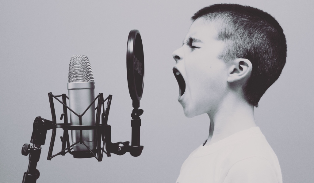Мальчик кричит в студийный микрофон на сером фоне 