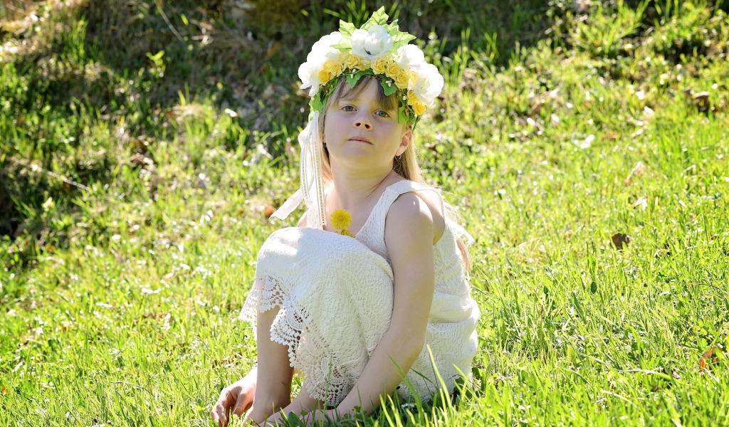 Маленькая девочка с венком на голове сидит на зеленой траве