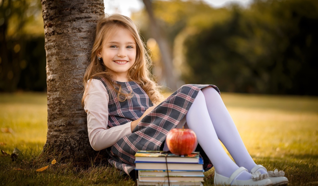 Маленькая девочка с книгами и яблоком сидит под деревом 