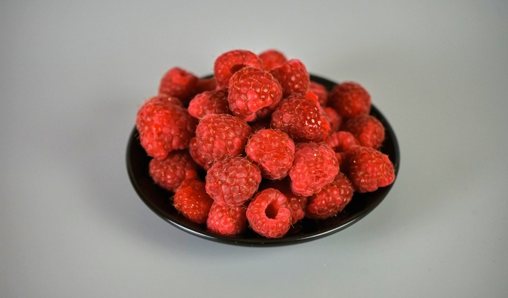 Спелая красная малина в черной тарелке на сером фоне