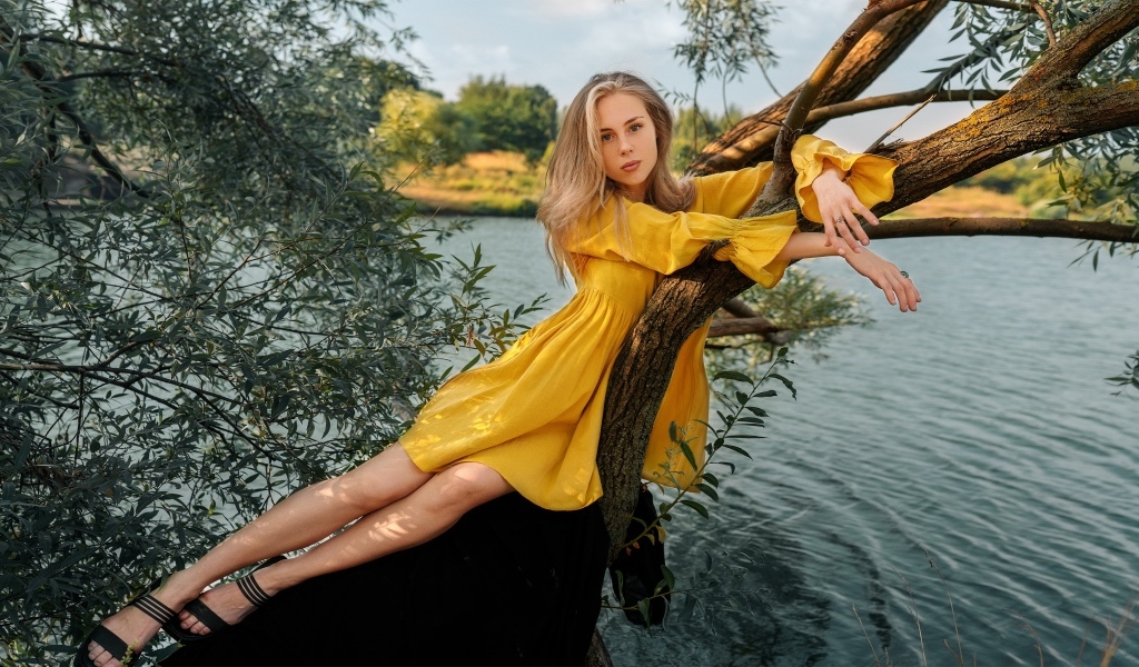 Красивая девушка в желтом платье лежит на дереве у воды