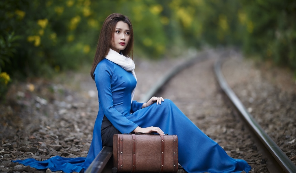 Красивая девушка азиатка сидит на рельсах с чемоданом