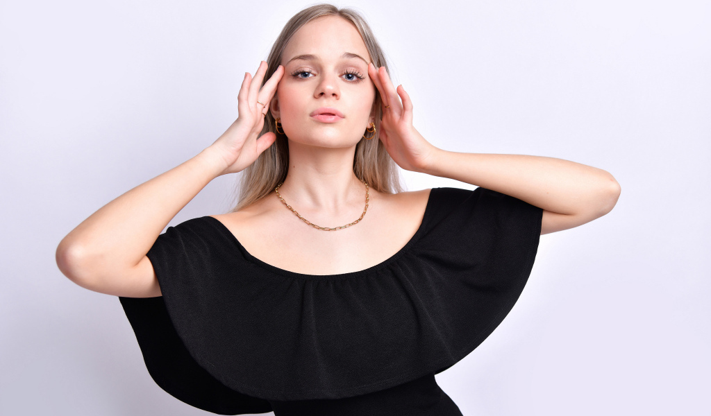 Девушка в черном платье с руками у головы на белом фоне