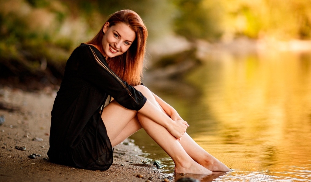 Улыбающаяся девушка в черном платье сидит у воды