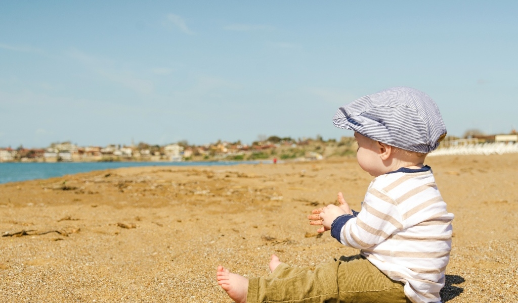 Маленький мальчик в кепке сидит на песке у воды
