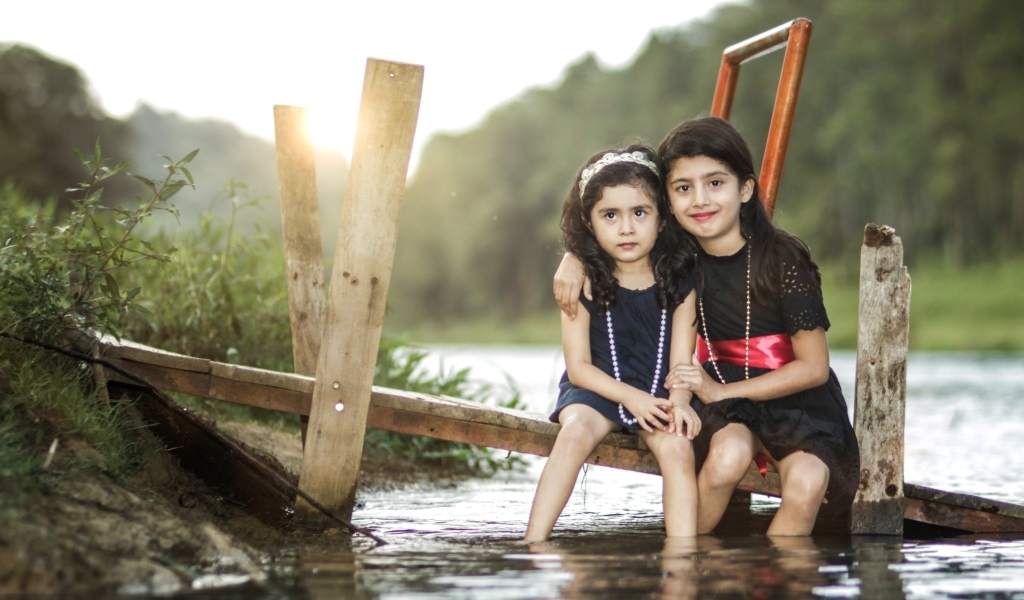 Две девочки сидят на мосту у реки