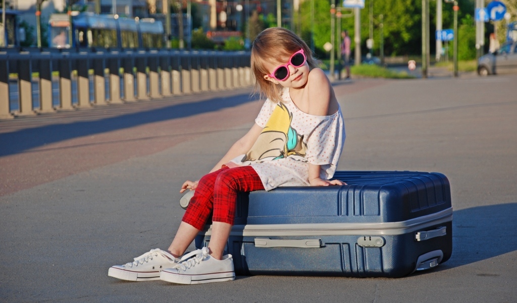 Девочка в солнечных очках сидит на чемодане на дороге