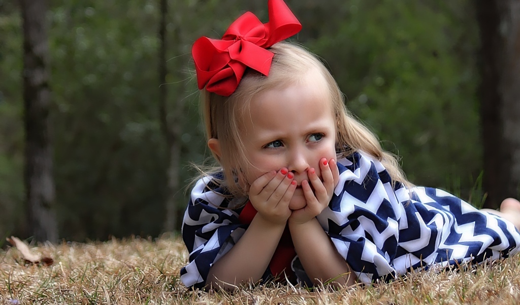 Маленькая девочка с красным бантом на голове лежит на траве