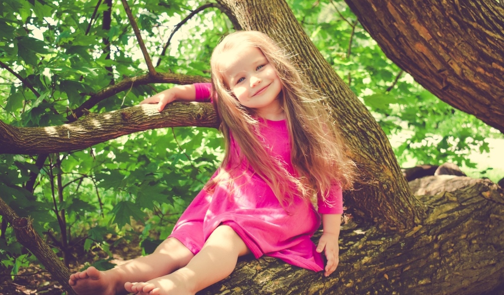 Милая девочка в розовом платье сидит на ветке дерева