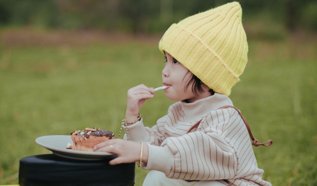 Маленькая девочка в желтой шапке ест пирожное