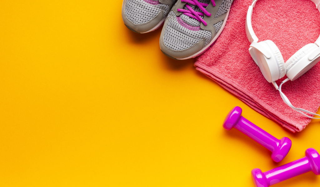 Розовые гантели, наушники, кроссовки и полотенце на желтом фоне