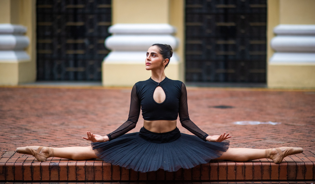 Девушка балерина в черном наряде сидит на шпагате