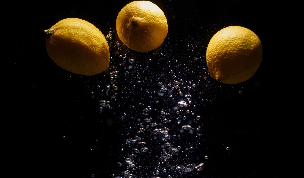 Три желтых лимона с пузырями в воде на черном фоне