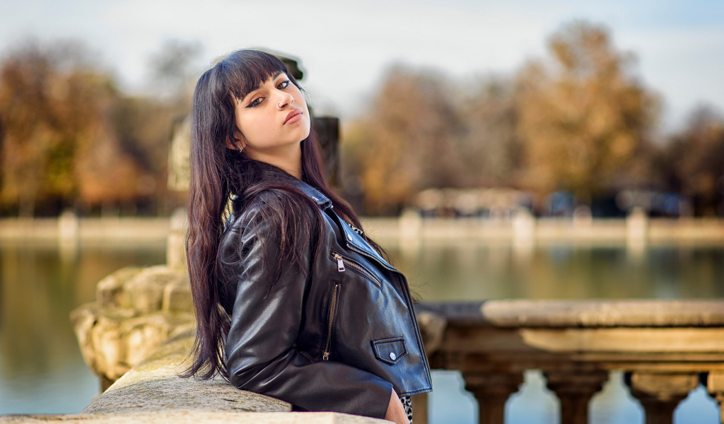 Девушка в черной куртке стоит на мосту у воды