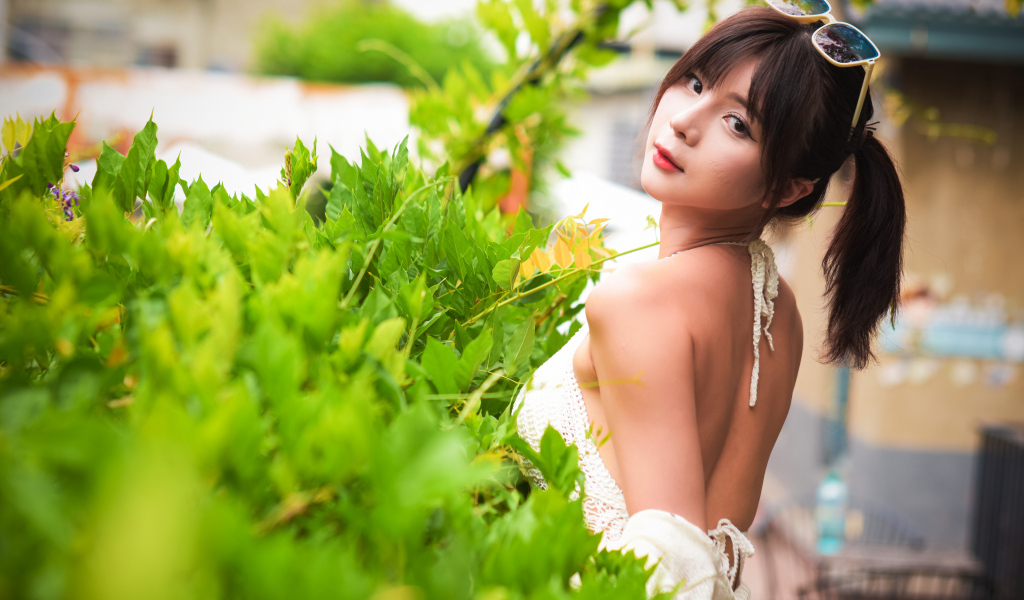 Девушка азиатка с очками на голове стоит у куста с зелеными листьями