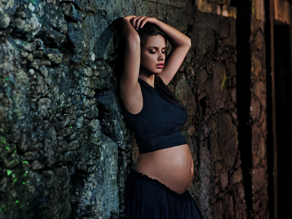 Беременная девушка в черном наряде у стены 