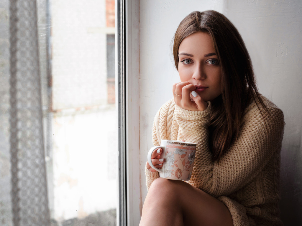 Молодая девушка в свитере сидит с чашкой на подоконнике