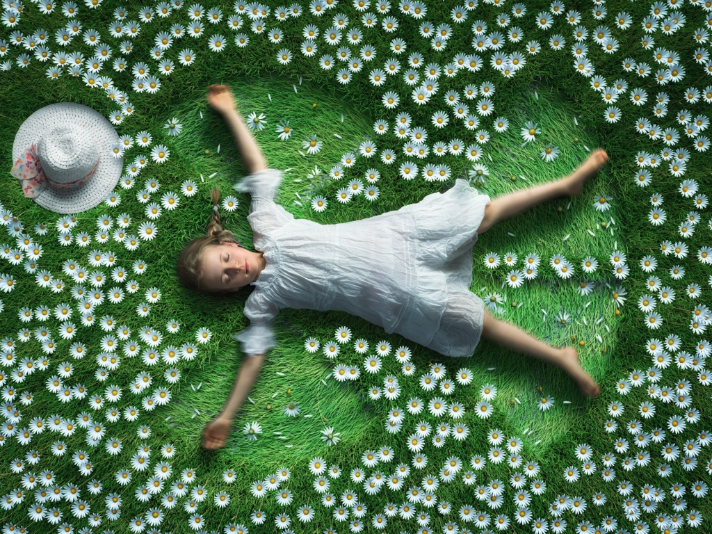 Маленькая девочка в белом платье лежит на зеленой траве с ромашками