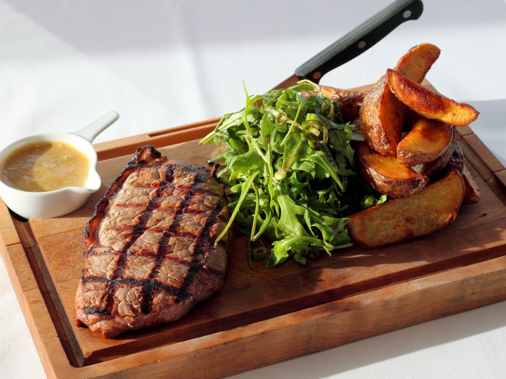 Стейк на доске с картофелем, соусом и рукколой