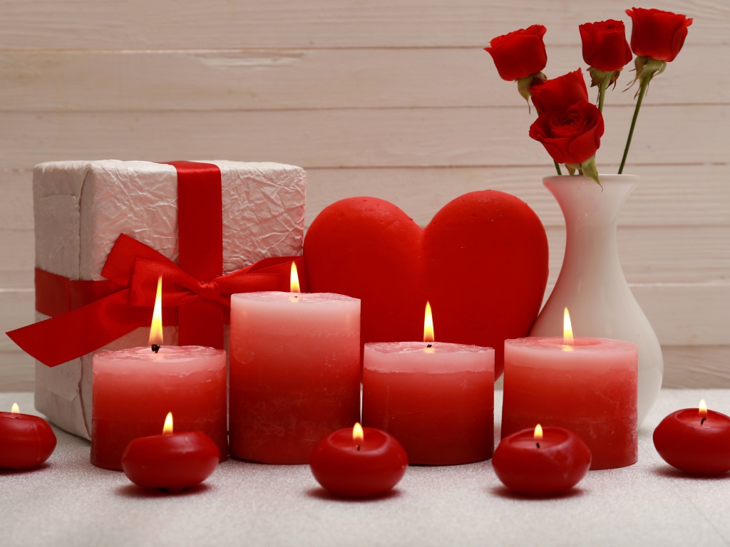 Зажженные красные свечи на столе с подарком, красным сердцем и цветами в вазе 