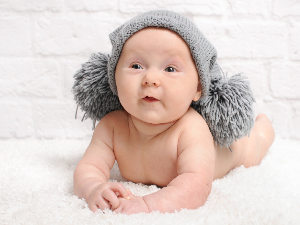 Улыбающийся грудной ребенок в серой шапке с бубонами