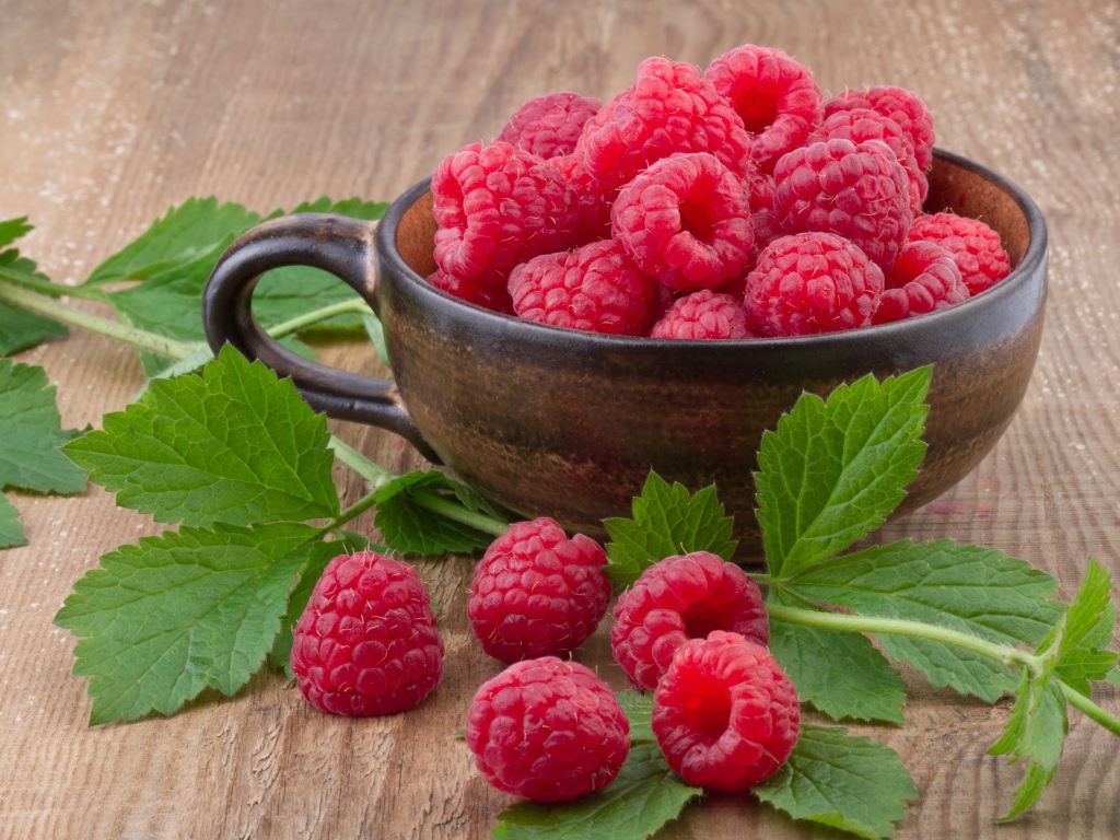 Чашка со свежей малиной на столе с зелеными листьями