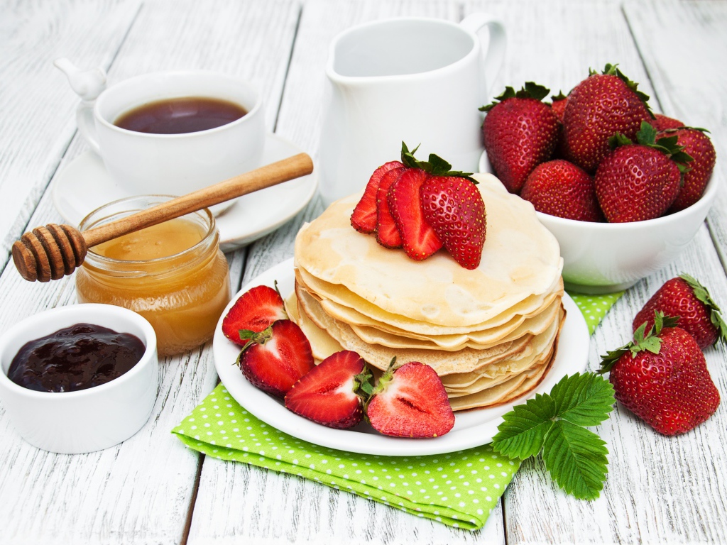 Тонкие блины на столе с ягодами клубники, медом, чаем и джемом