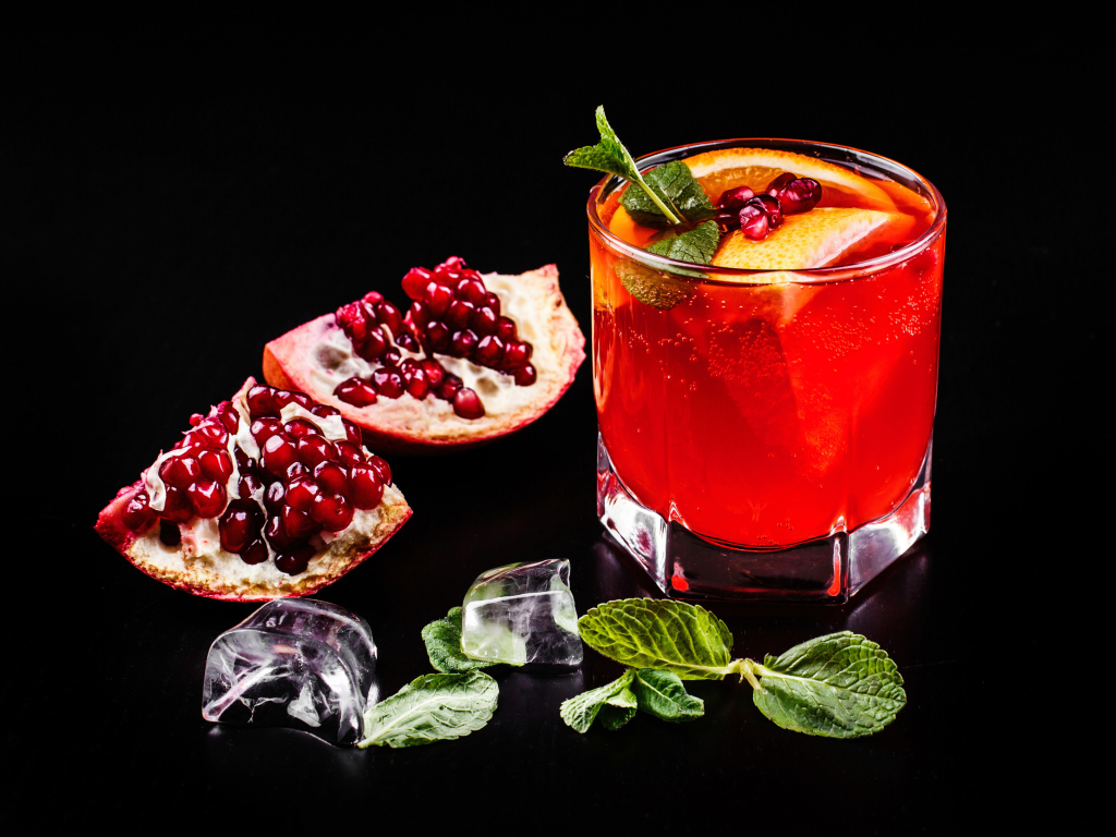 Алкогольный напиток в стакане на черном фоне с гранатом, кусочками льда и мятой