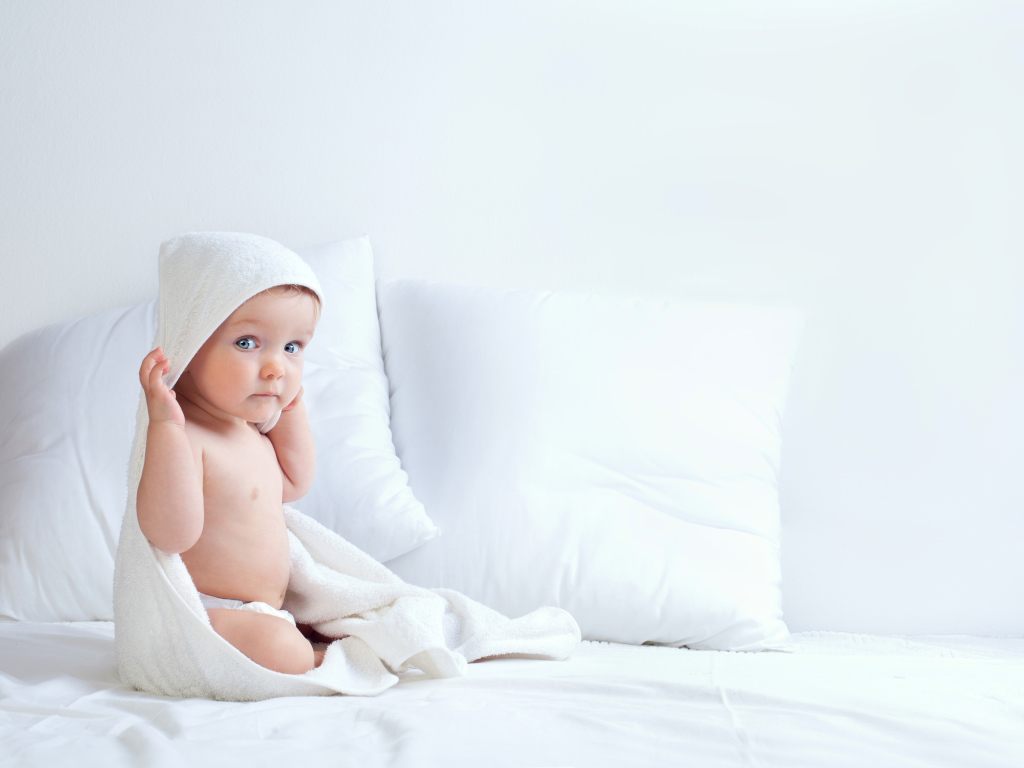 Маленький мальчик сидит в белом покрывале на кровати с подушками