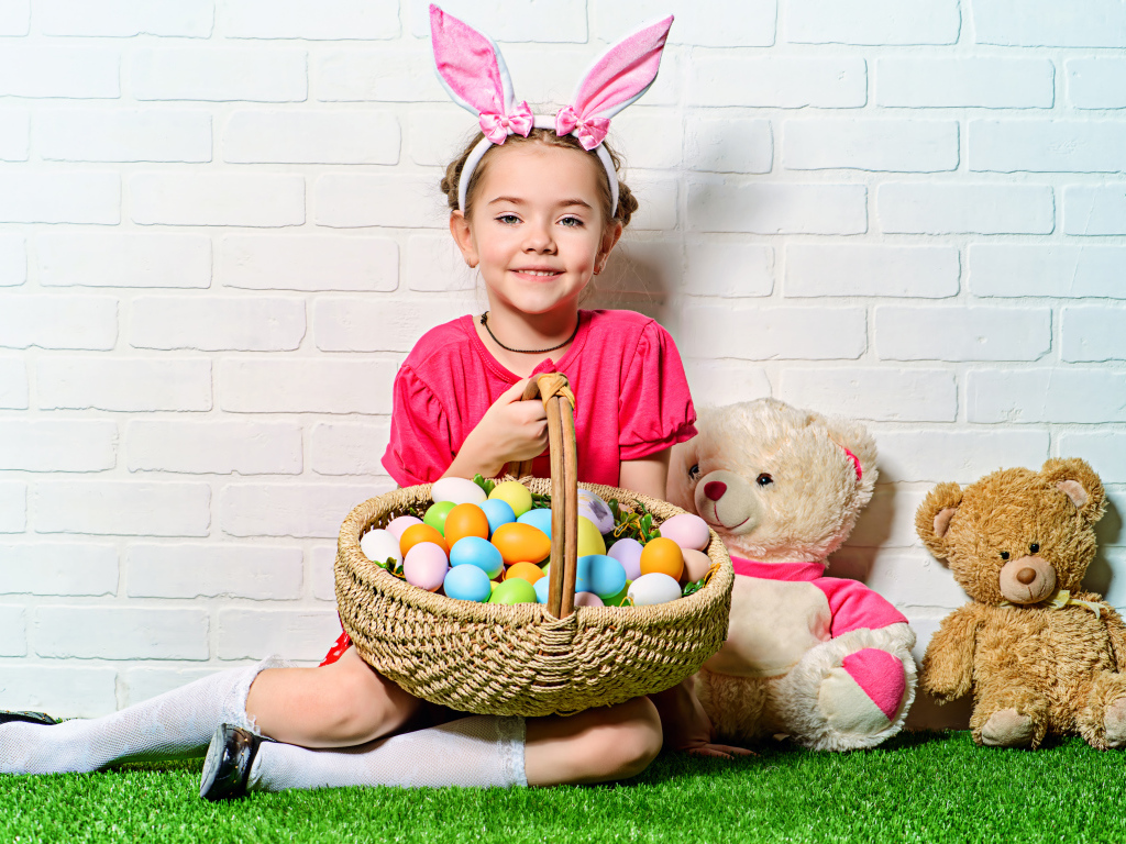 Улыбающаяся девочка сидит у стены с корзиной пасхальных яиц и игрушками