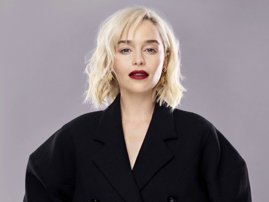 Яркая блондинка, актриса Эмилия Кларк в черном пальто на сером фоне